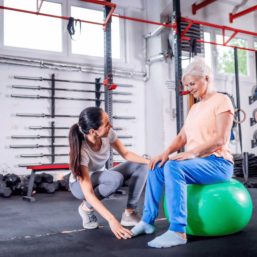 Therapeutin begleitet ältere Frau bei Rehaübung mit Gymnastikball im Trainingsbereich